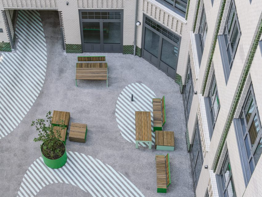 Courtyard with several wooden benches and tables on a grey floor with white striped oval surfaces, surrounded by light-coloured building facades with windows