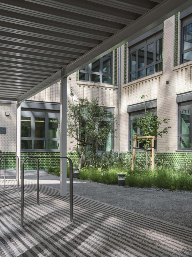 Inner courtyard with paved ground, young trees with supporting frames and a building with light-coloured brick facades and large windows