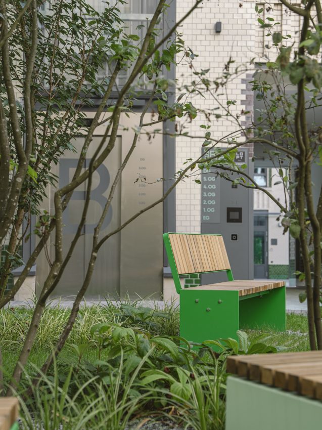 Green bench with wooden slats in a leafy courtyard in front of a building with light-coloured clinker bricks and lift signage