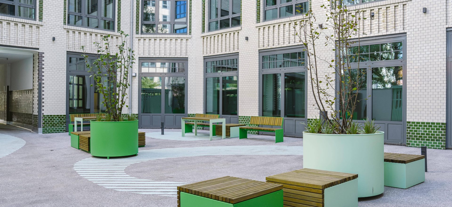 Inner courtyard with round planters in green and pastel colours, wooden seating areas and modern benches in front of a multi-storey building with many windows
