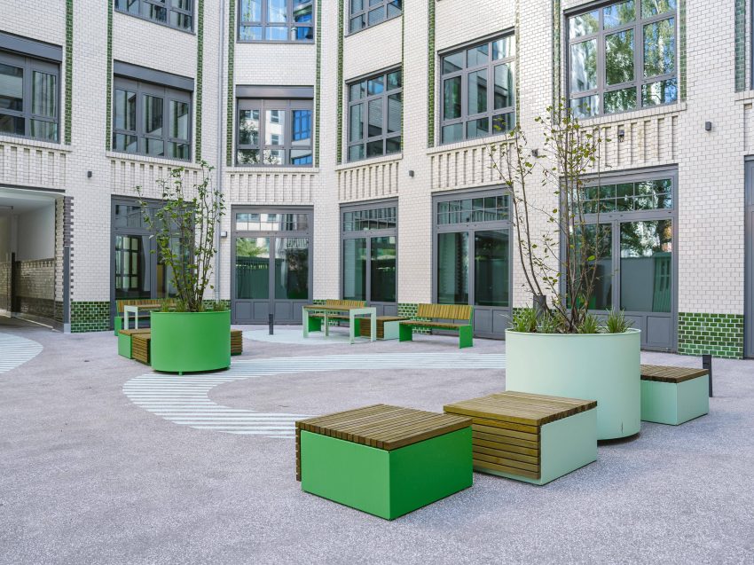 Inner courtyard with round planters in green and pastel colours, wooden seating areas and modern benches in front of a multi-storey building with many windows