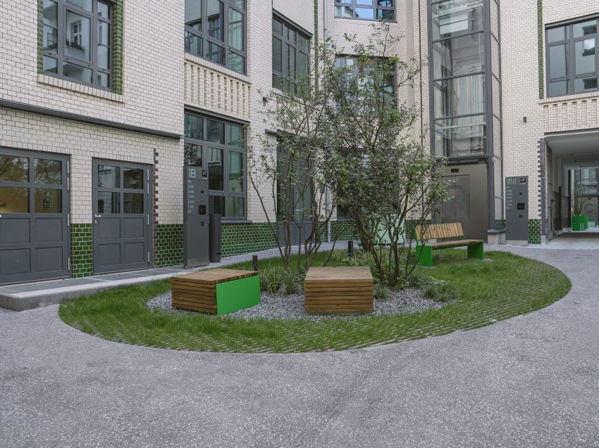 Innenhof mit Sitzbänken aus Holz und grünen Metallteilen, Büschen in Kiesbett, umgeben von mehrstöckigem Gebäude mit weißen Fassaden und großen Fenstern